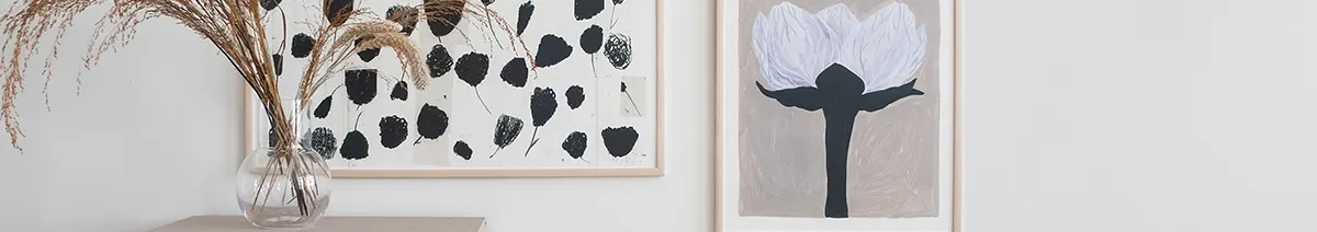 Två inramade konstverk och en vas med torkat gräs. Vänster bild har svarta, abstrakta former; höger har en stor lila blomma.