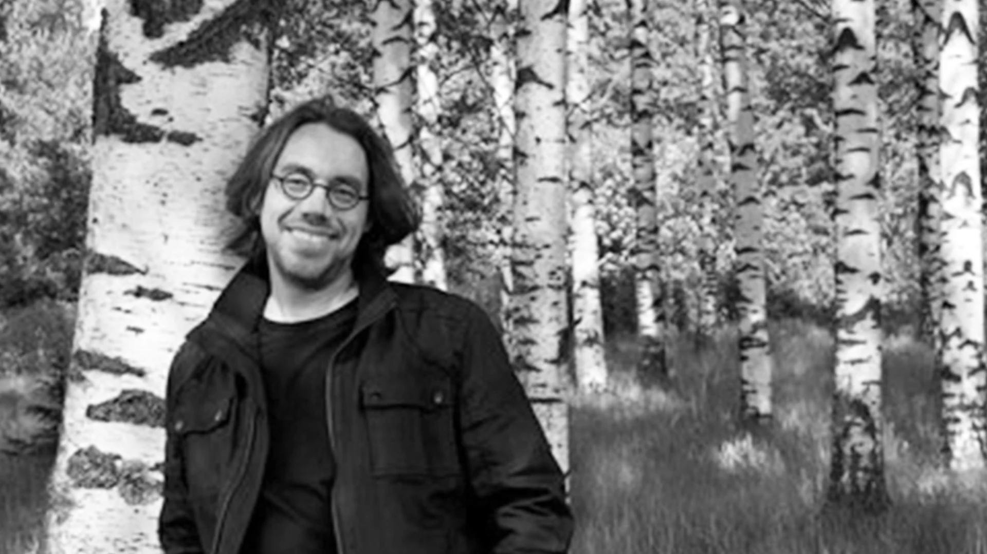 En leende man med glasögon och långt hår lutar sig mot en björk, med flera björkträd i bakgrunden.