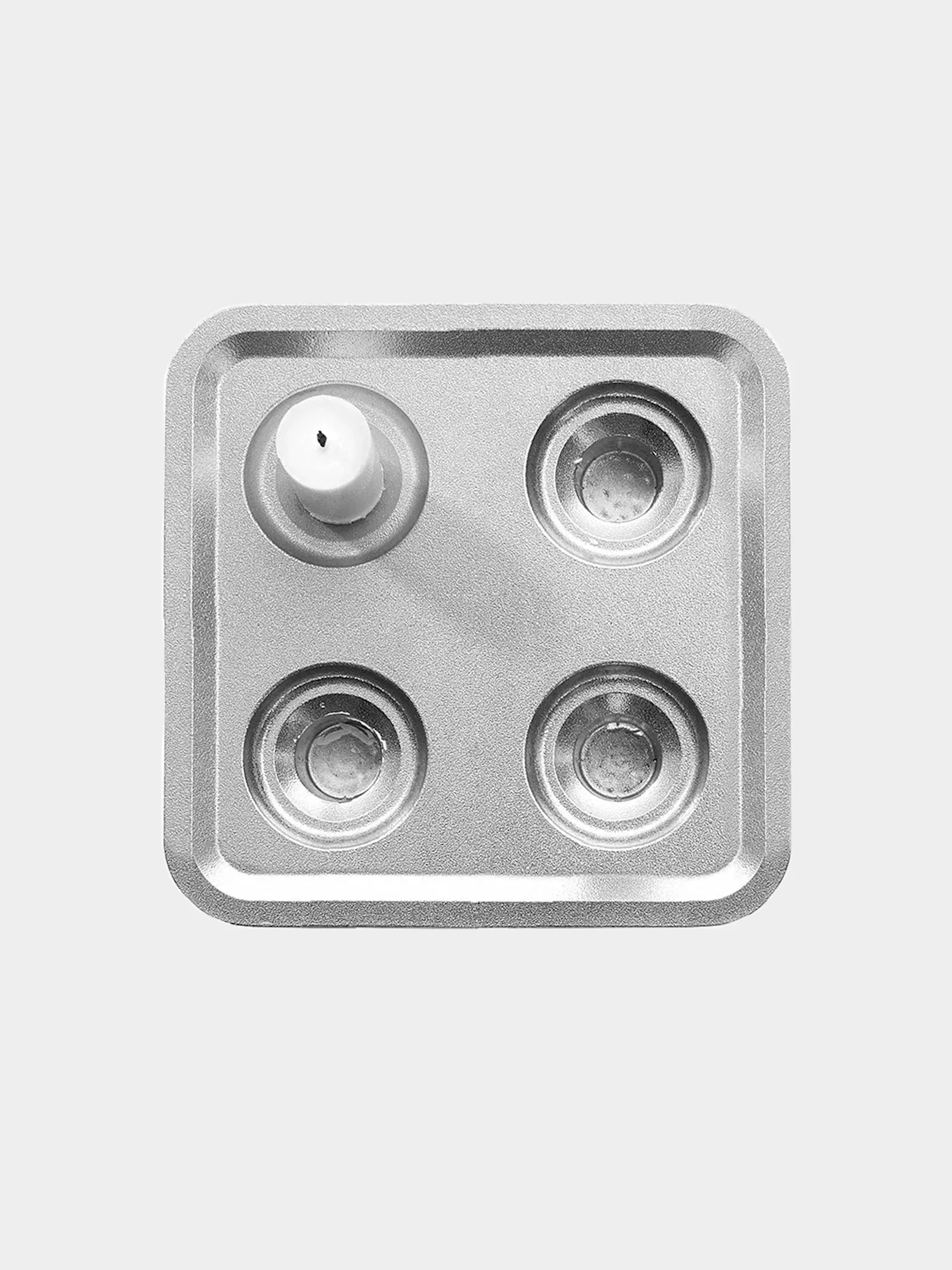 Fyrkantig ljusstake i silver med ett vitt ljus i övre vänstra av fyra fördjupningar.