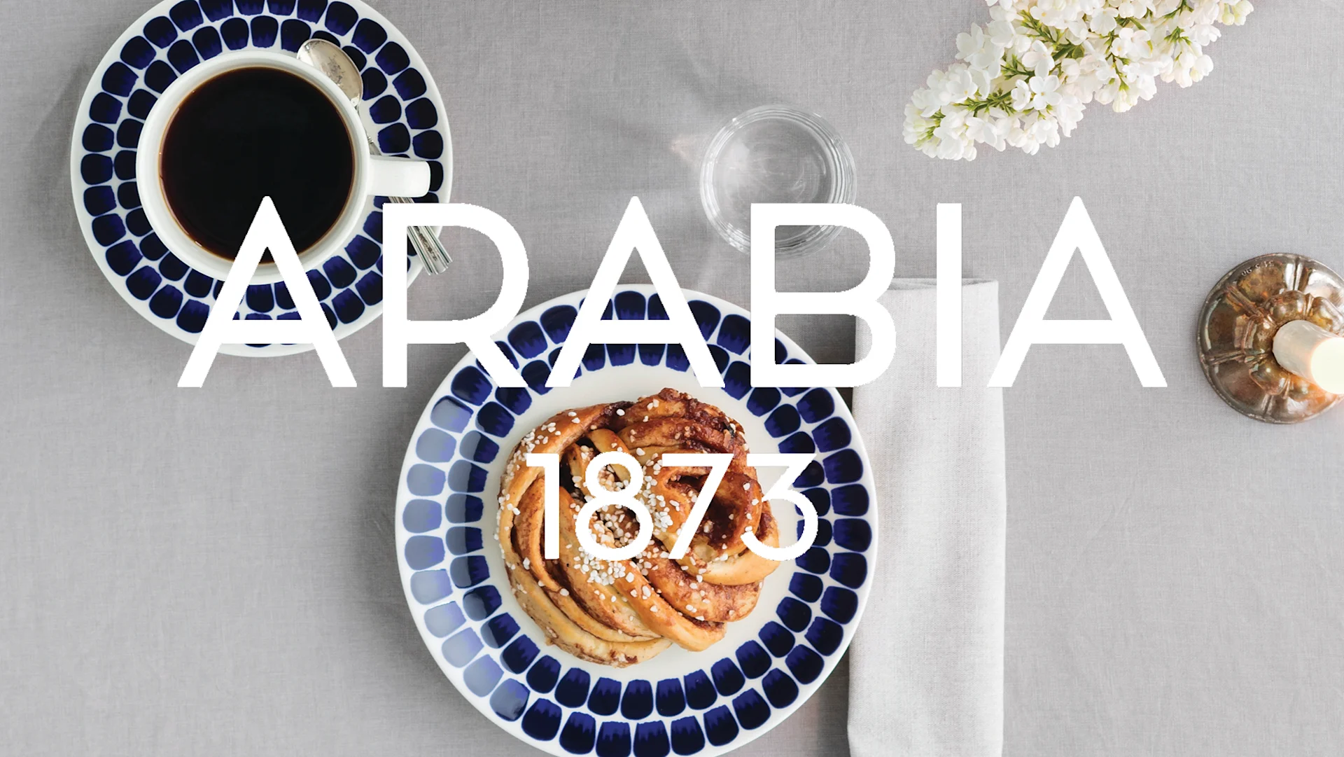 ARABIA 1873 över ett bord med kaffe, kanelbulle på blåvita serviser, vit syren och ljus.