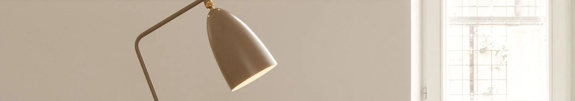 Närbild av en modern gråbrun lampa med konisk skärm som avger ljus, bredvid ett fönster med rutnät.