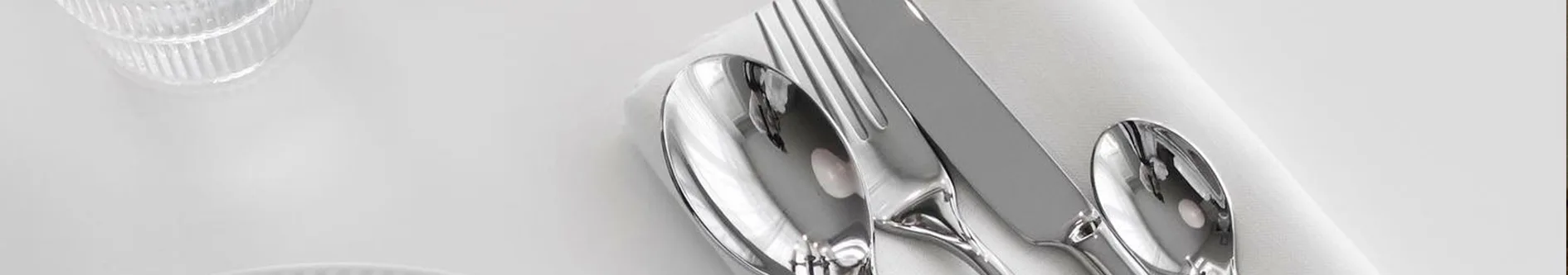 Glänsande silverfärgat bestick (gaffel, sked, kniv) på en vit tygservett med ett klart glas i bakgrunden.