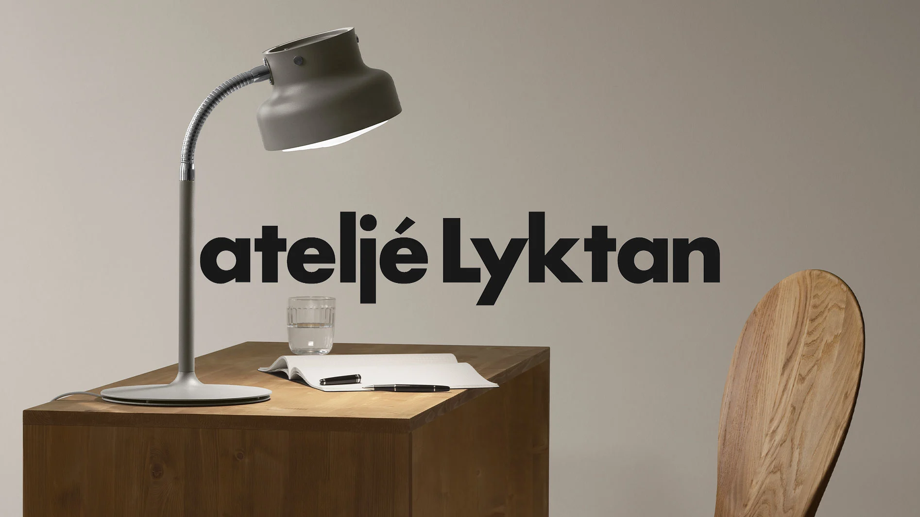 Grå bordslampa på ett träbord med anteckningsbok, pennor och ett glas vatten. ateljé Lyktan-logotypen är centrerad.