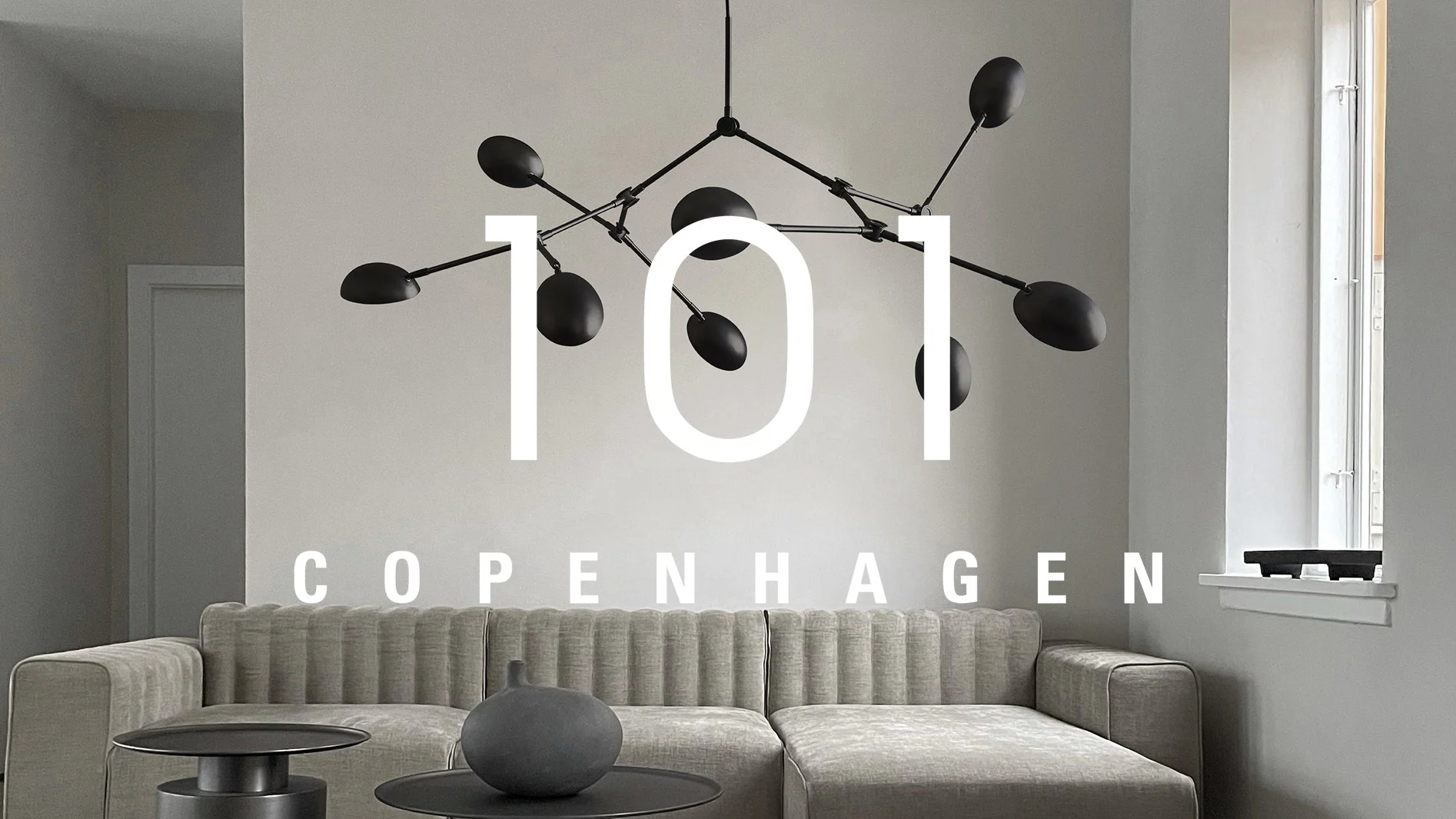 Ljusgrå ribbad sektionssoffa, svarta sidobord, mörkgrå vas och svart modern taklampa i ett vardagsrum. Text: "101 COPENHAGEN".