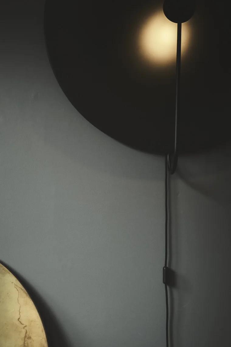 Mörk, cirkulär vägglampa med varmt ljus mot en grå vägg. En sladd med strömbrytare hänger ner.