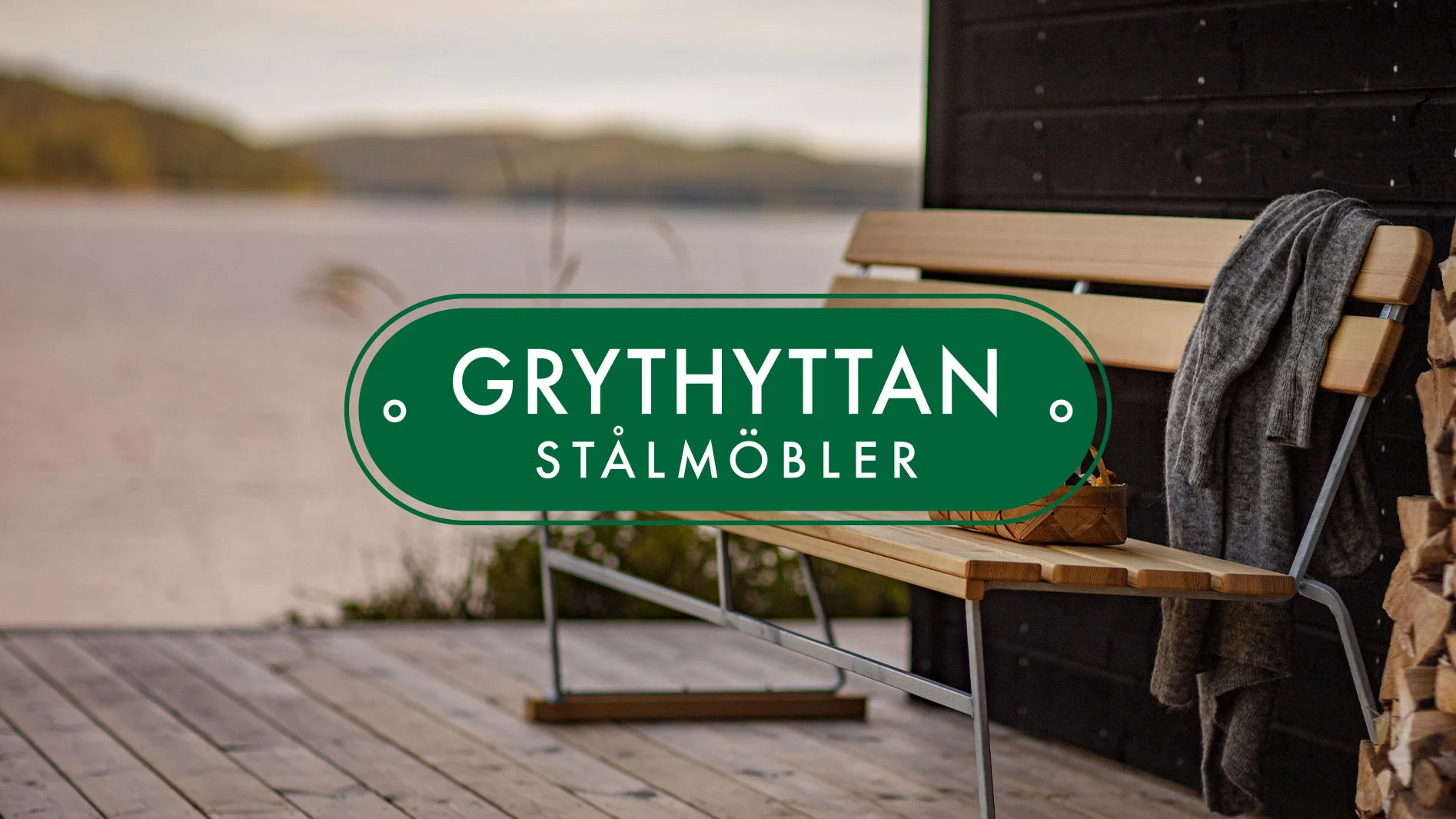 Träbänk med metallram, grå filt, vedstapel på trädäck vid sjö. Grythyttan Stålmöbler-logotyp.