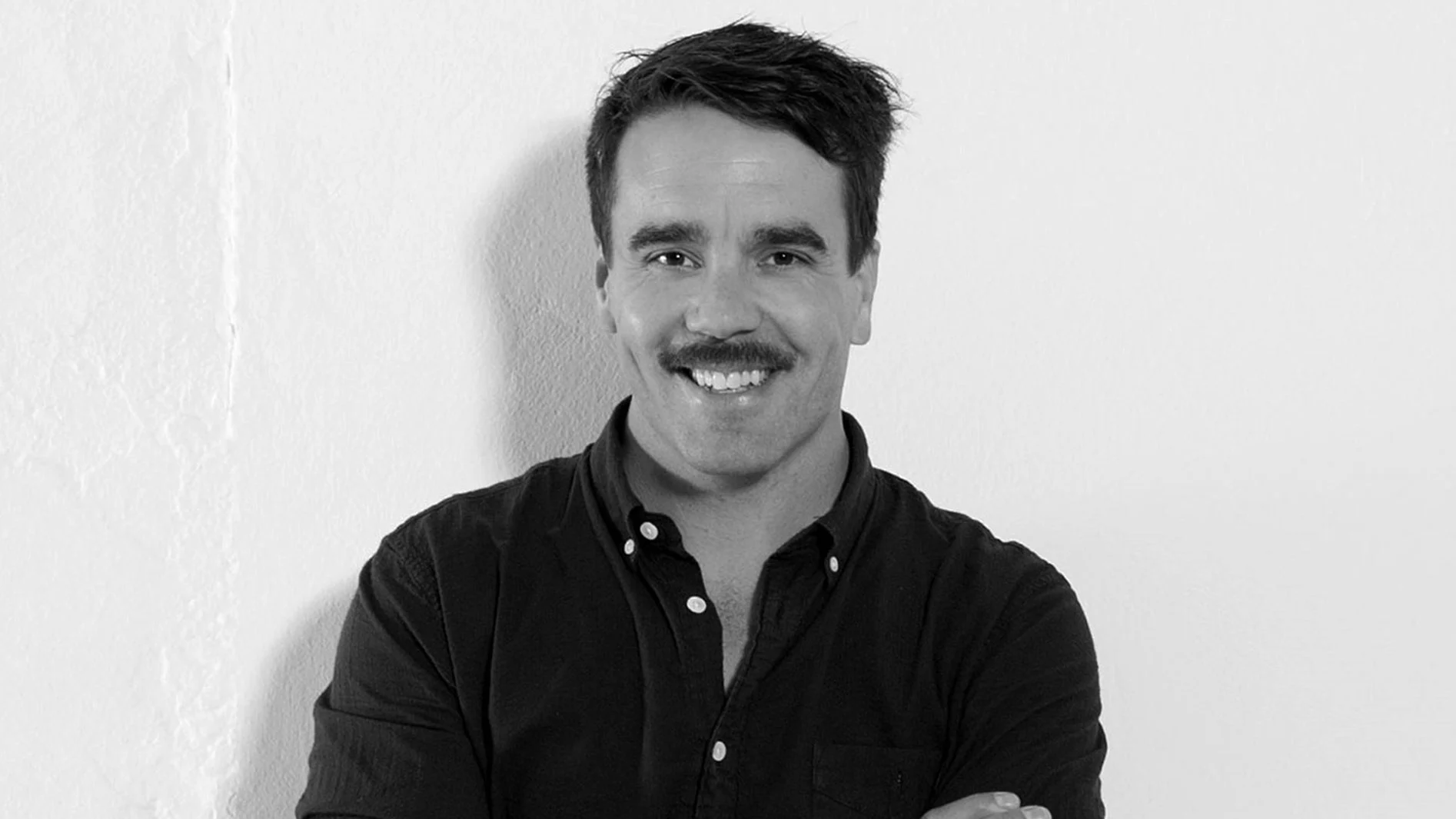 Svartvitt porträtt av en leende man med mustasch och mörk skjorta.