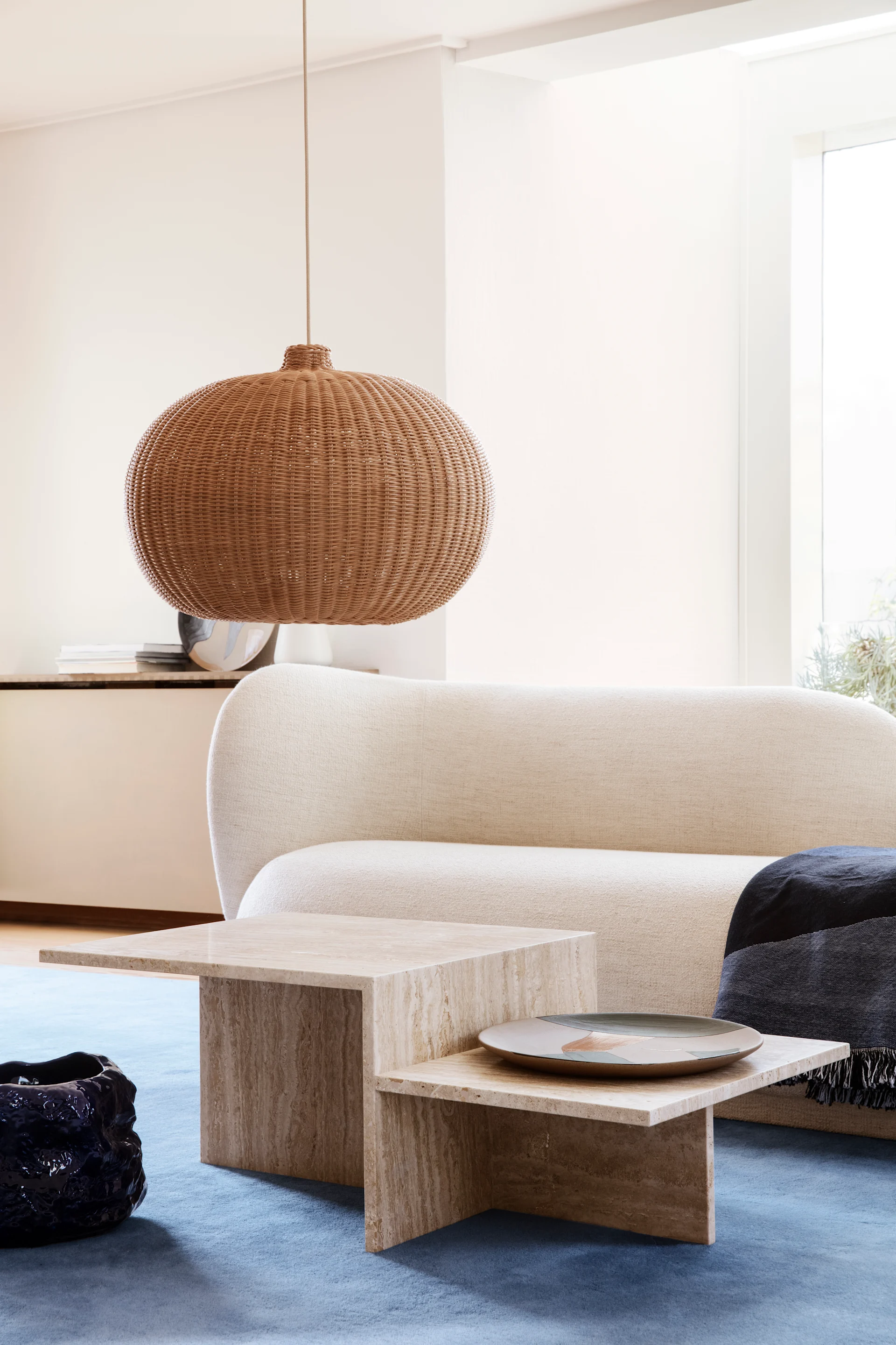 Så väljer du rätt soffbord, bild som visar Distinct soffbord i stenen travertine från Ferm Living, placerat på en blå matta framför en vit soffa, över bordet hänger en lampa i rotting.