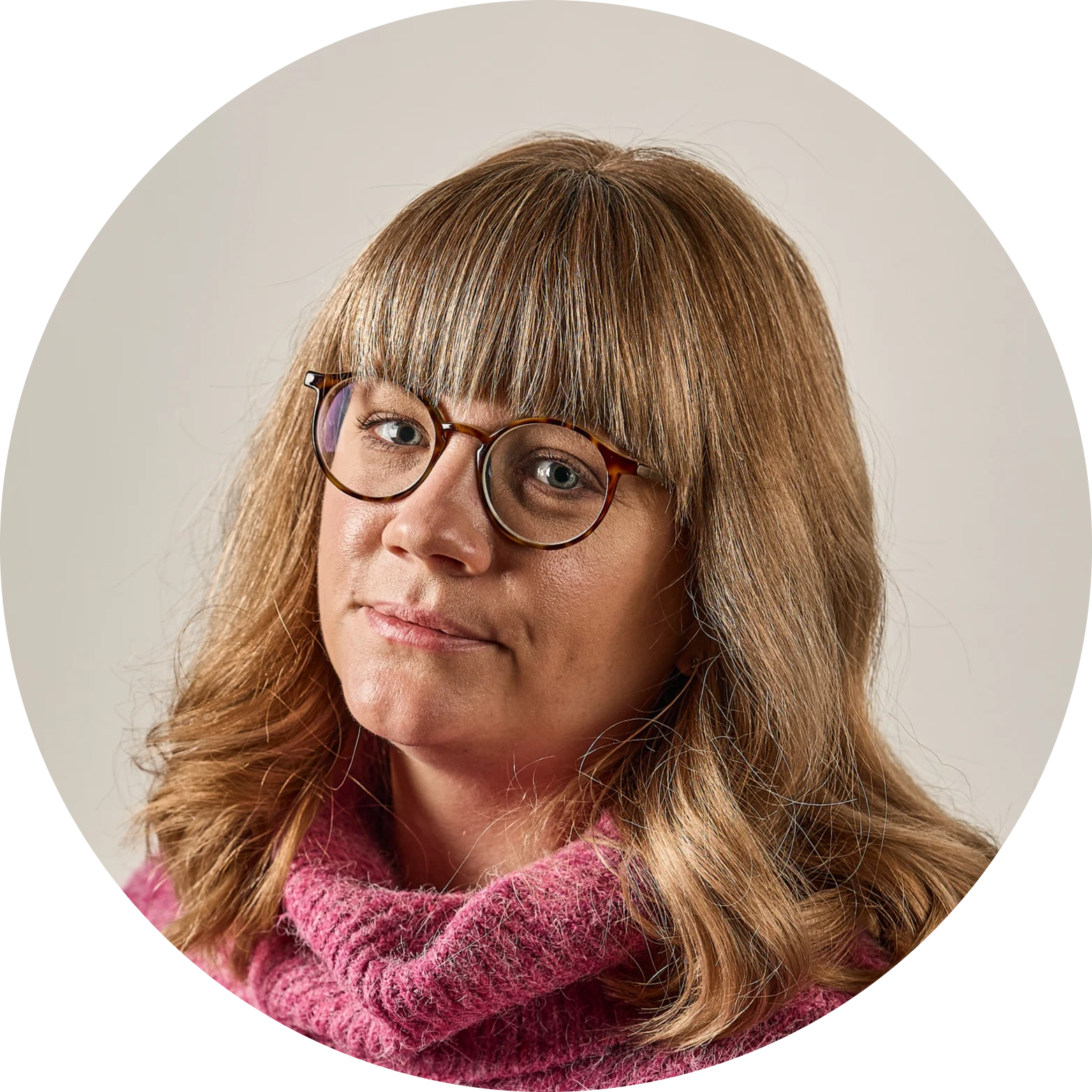 En kvinna med blont hår, lugg och glasögon, iklädd en rosa tröja, tittar direkt in i kameran.