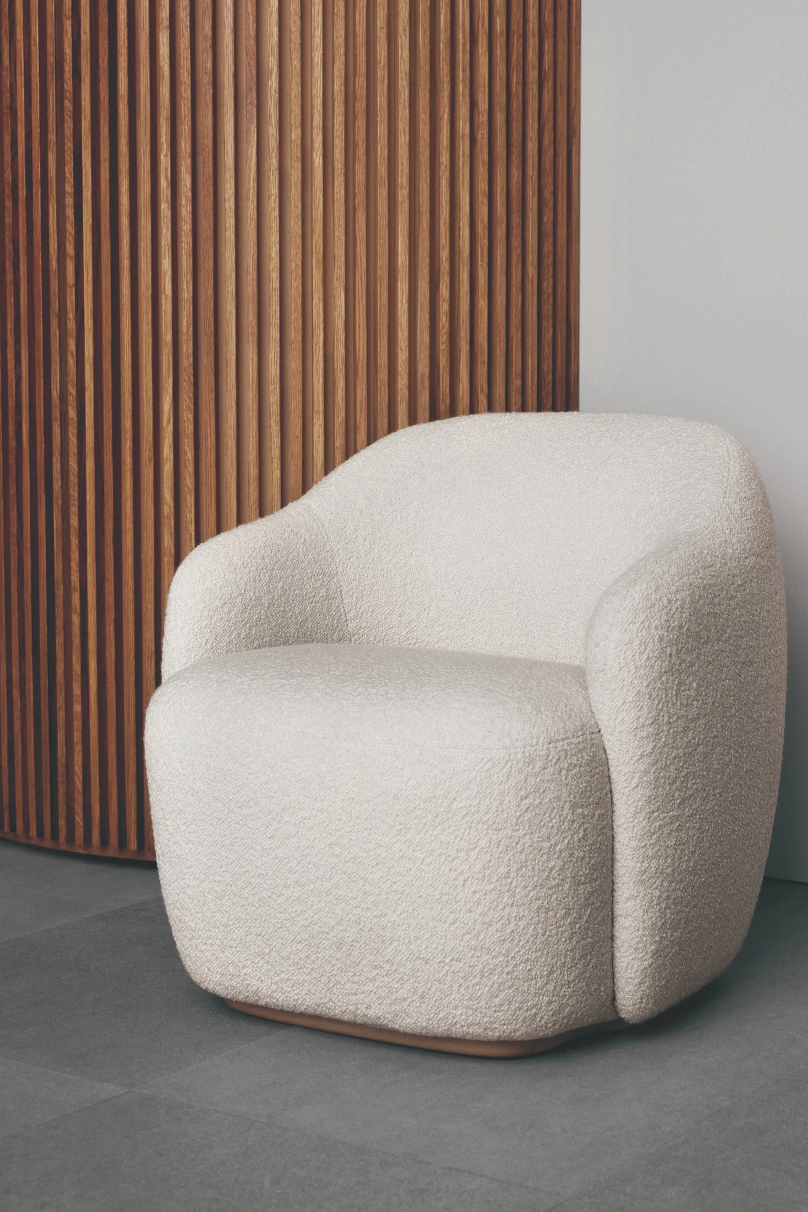 Krämfärgad bouclé-fåtölj med böjd design och träbotten, mot en räfflad träpanelvägg och grått golv.