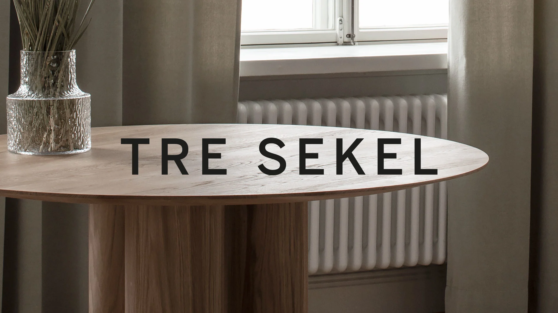 Runt träbord med texten "TRE SEKEL", vas med torkade växter, och fönster i bakgrunden.