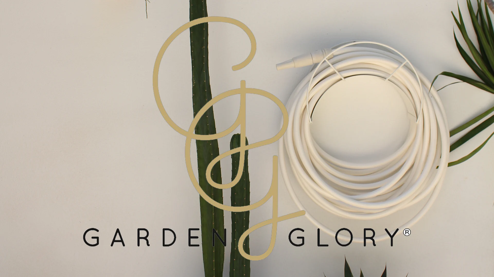 En upprullad vit trädgårdsslang med munstycke på ljus bakgrund med gröna växter och Garden Glory-logotypen.