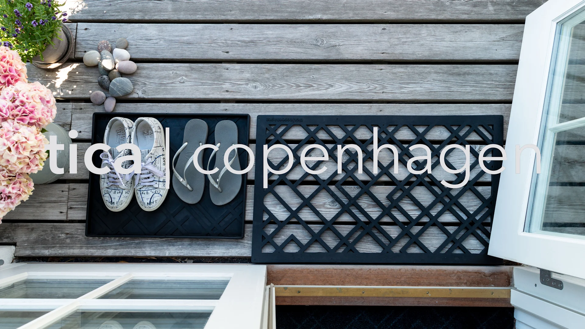 Översiktsbild av ett trägolv med krukväxter, ett par vita sneakers och grå flip-flops på två svarta mattor, bredvid ett öppet vitt fönster.