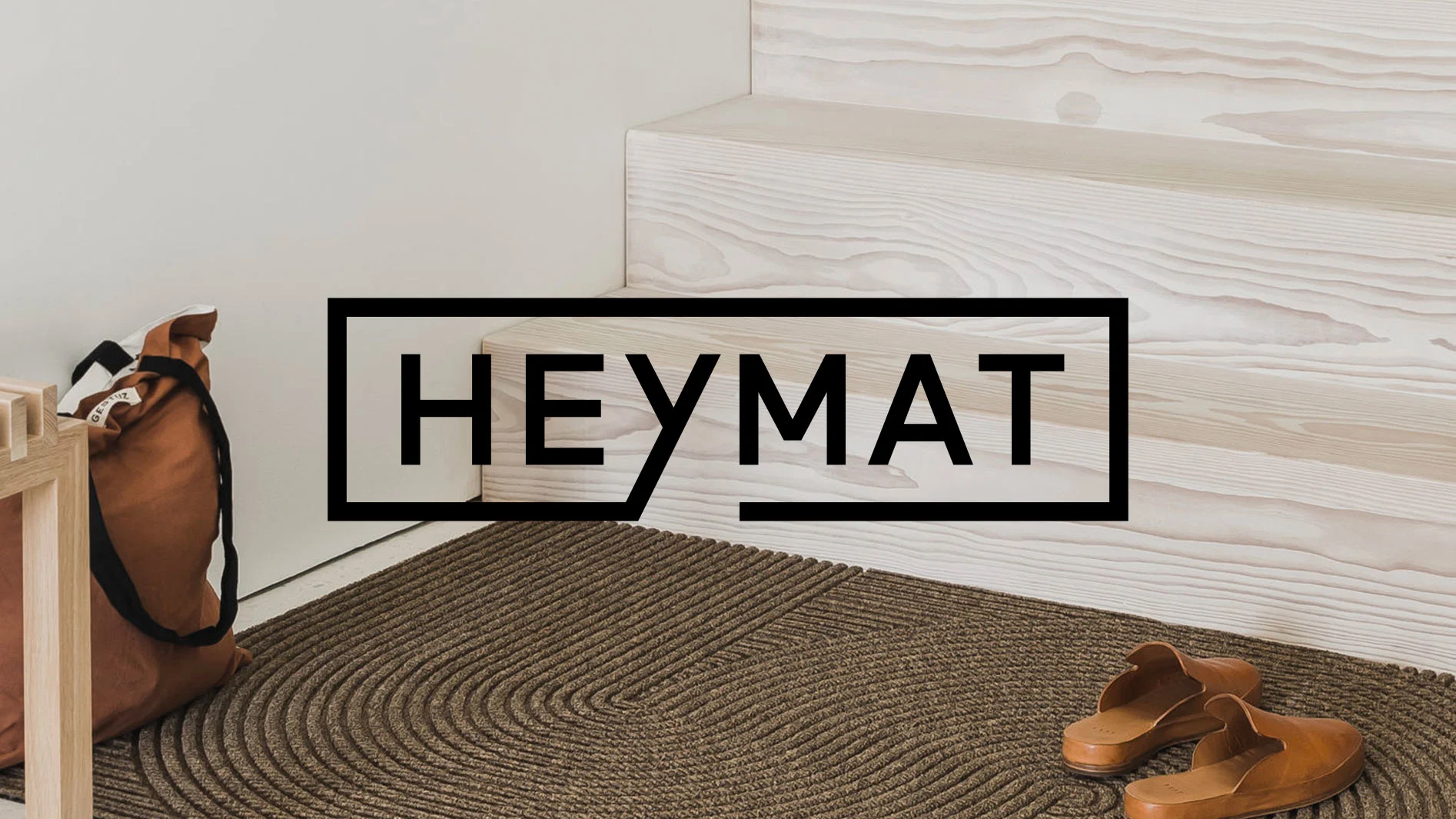Brun flätad dörrmatta vid en ljus trappa, med bruna muleskor och en tygväska. HEYMAT-logotypen.