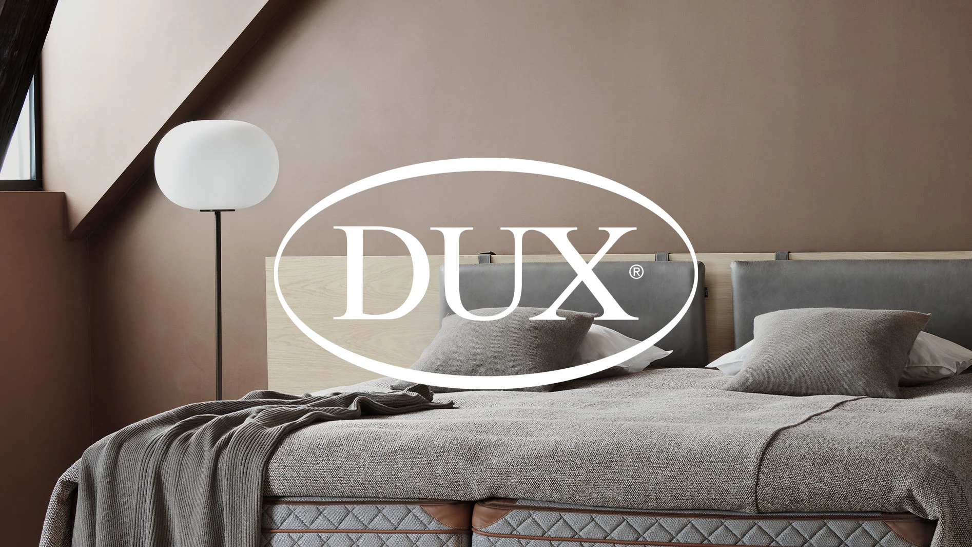 Sovrum med DUX-säng, grå sängkläder, ljusbruna väggar och vit golvlampa. DUX-logotyp i förgrunden.