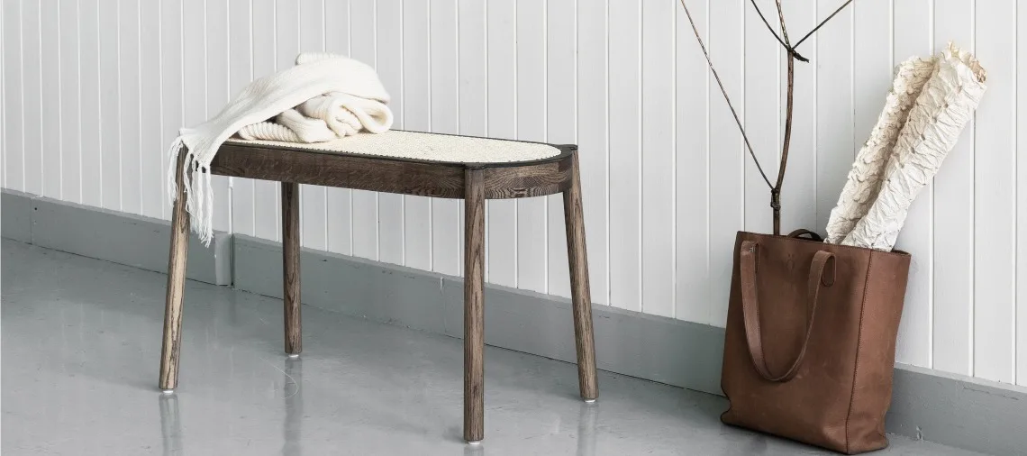 Mörk träbänk med flätad sits, vit filt och brun läderväska med en rullad pläd.